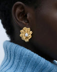 A close-up of a person wearing For Art's Sake® The Shell Earrings—large gold flower-shaped earrings with a freshwater pearl center—paired with a light blue ribbed turtleneck, showcasing an elegant coastal jewelry style.