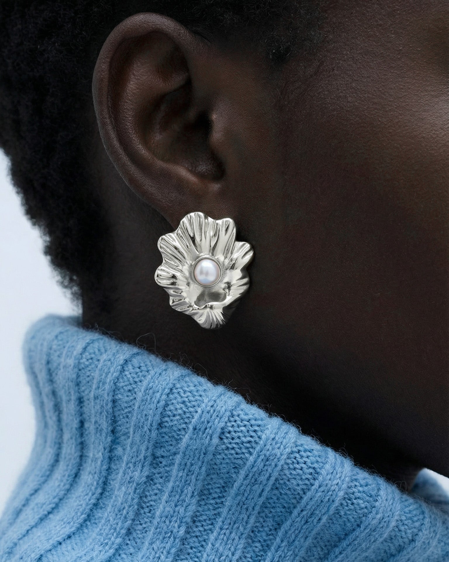 A close-up of someone wearing The Shell Earrings by For Art's Sake®—silver flower-shaped with a freshwater pearl center—paired with short curly hair and a light blue ribbed turtleneck for an everyday look.