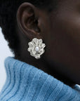 A close-up of someone wearing The Shell Earrings by For Art's Sake®—silver flower-shaped with a freshwater pearl center—paired with short curly hair and a light blue ribbed turtleneck for an everyday look.