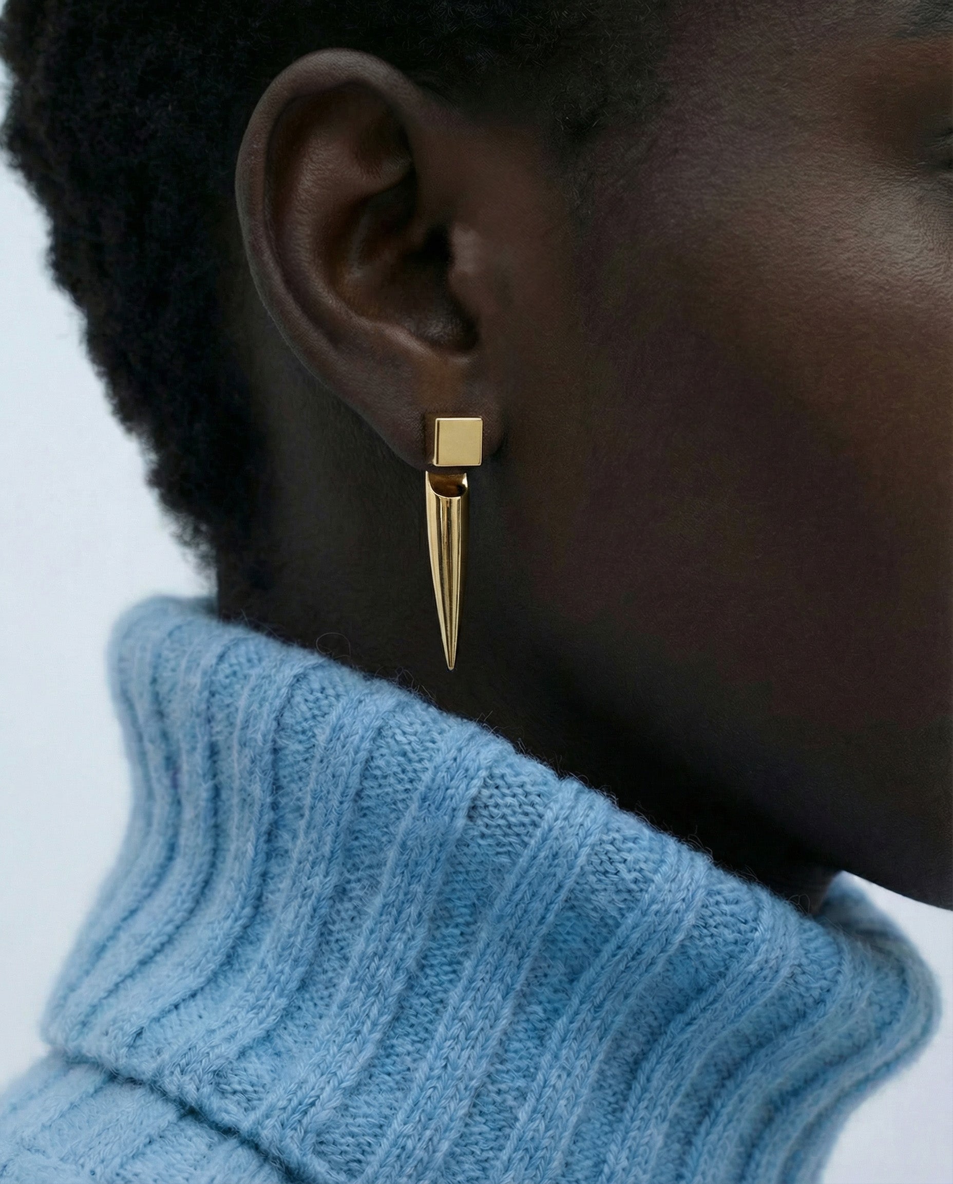 Close-up of a person wearing For Art's Sake® The Droplets Earrings—gold geometric earrings with a square stud and long drop—styled with short hair and a light blue ribbed turtleneck.