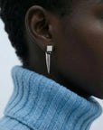 Close-up of a person's ear wearing The Droplets Earrings by For Art's Sake®—a silver geometric design with a square stud and long, pointed pendant. Paired with a light blue ribbed turtleneck, the lightweight jewellery stands out.