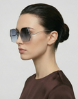 A woman with dark hair in a neat bun models For Art's Sake® Generation oversized geometric sunglasses and a dark brown top, shown in profile against a plain light background.