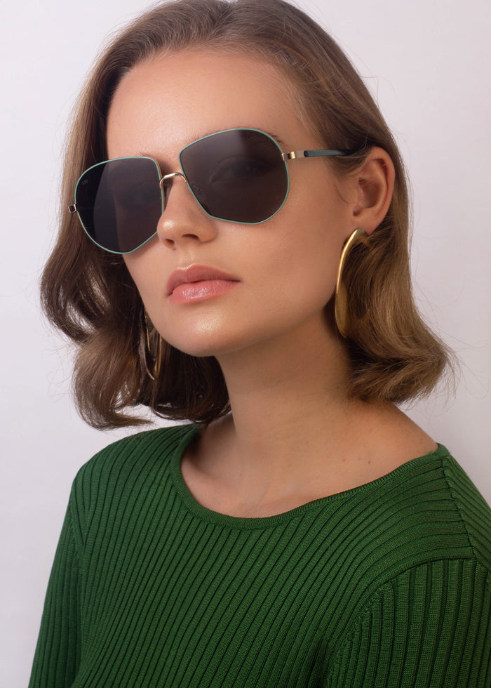 A woman with wavy light brown hair is wearing For Art's Sake® Explorer sunglasses that offer 100% UV protection, large hoop earrings with 18-karat gold plating, and a green ribbed top. She is posing against a plain background, looking slightly to the side with a neutral expression.