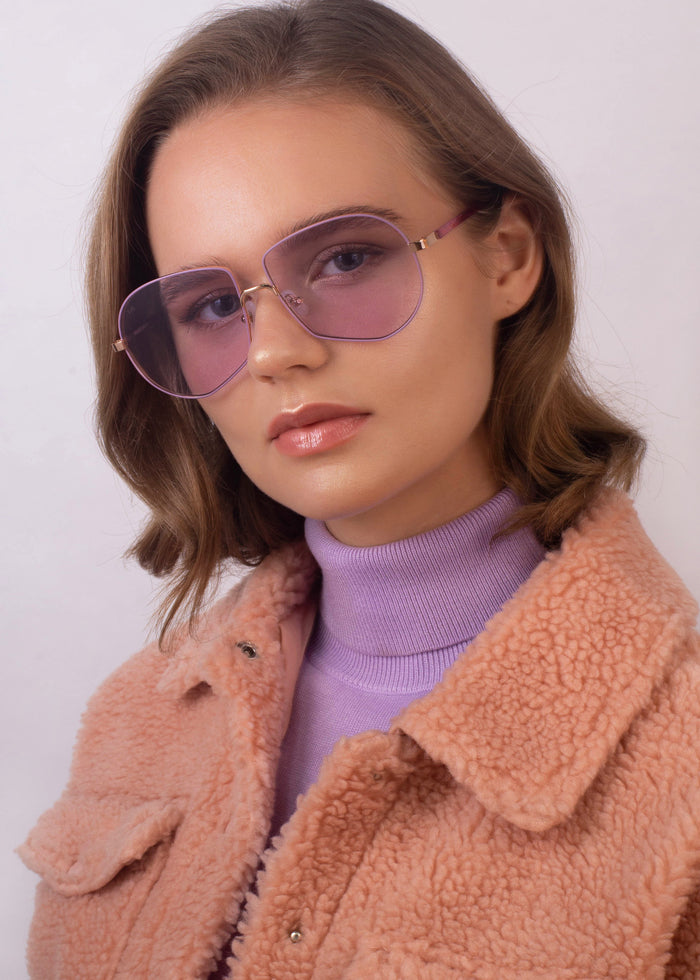 An individual with short, wavy hair sports the For Art's Sake® Explorer UV-protected aviator sunglasses, a lilac turtleneck, and a peach faux-shearling jacket. They gaze straight ahead with a neutral expression against a plain background.