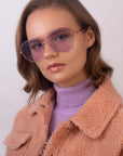 An individual with short, wavy hair sports the For Art's Sake® Explorer UV-protected aviator sunglasses, a lilac turtleneck, and a peach faux-shearling jacket. They gaze straight ahead with a neutral expression against a plain background.