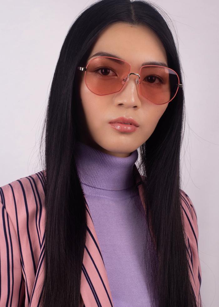A woman with long straight black hair wears For Art's Sake® Explorer pink-tinted aviator sunglasses (worn by Beyoncé), paired with a lilac turtleneck and pink pinstripe blazer, posing confidently against a light background.