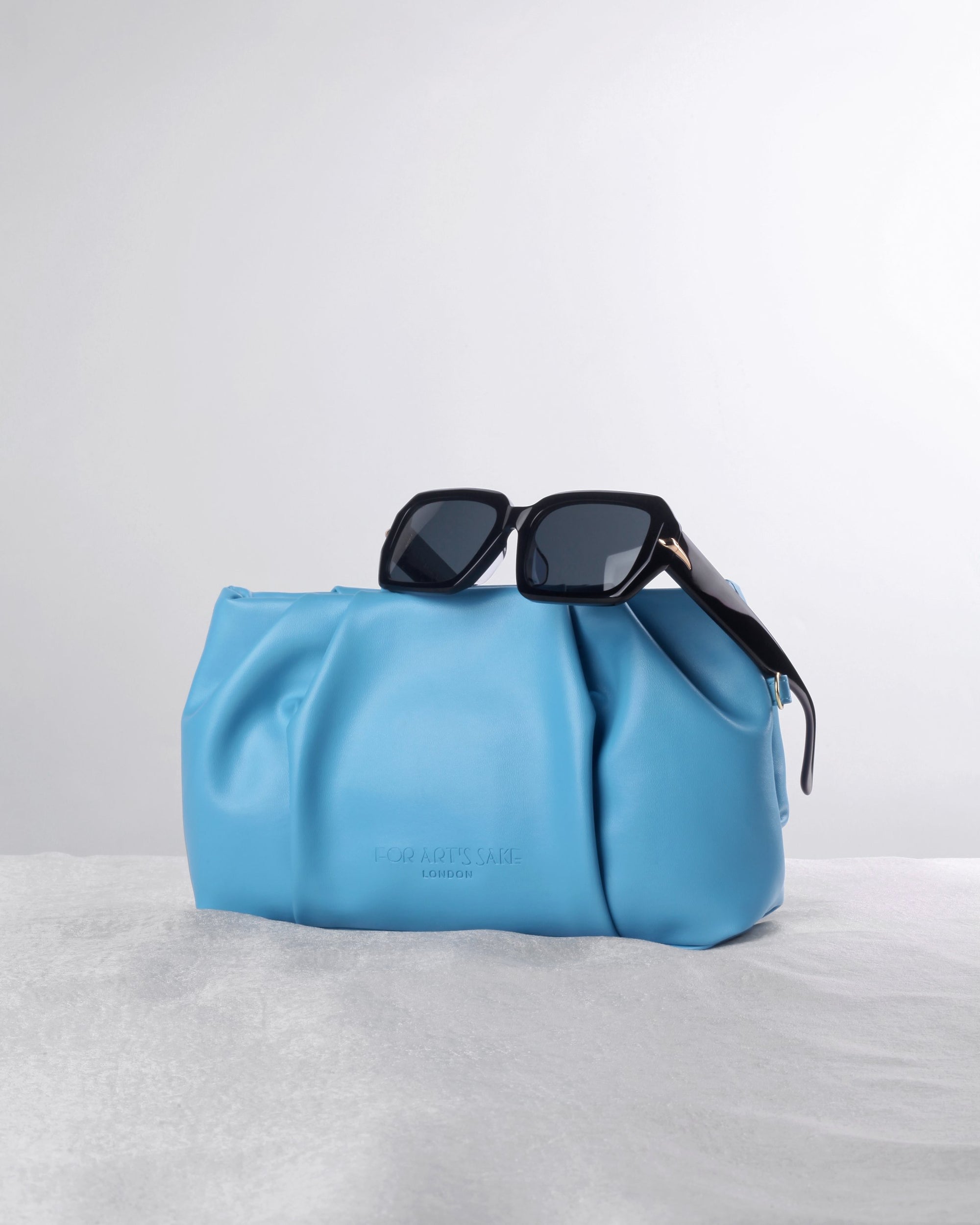 A pair of black sunglasses from FOR ART'S SAKE® sits atop the Soft packaging clutch in light blue leather with detachable chains, displayed on a textured white surface against a plain, light background.