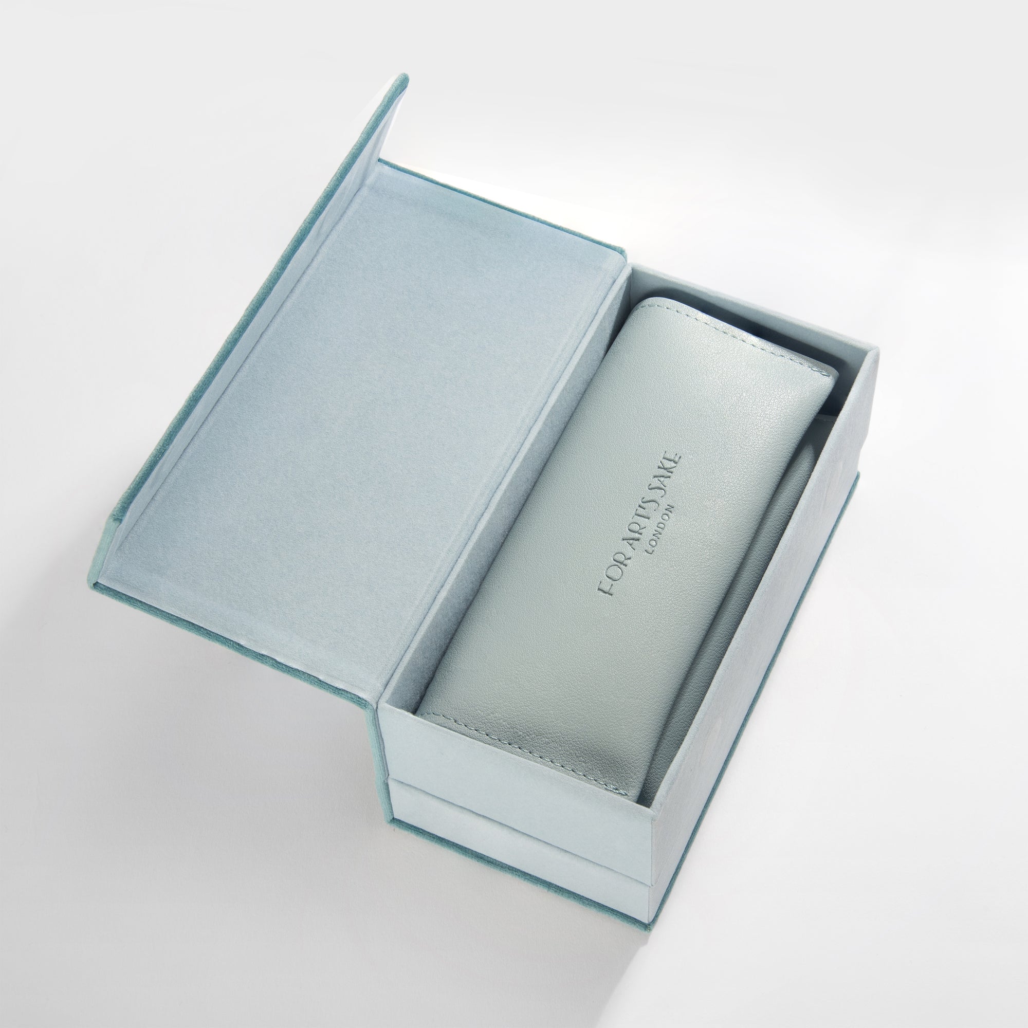 A pale blue box with a partially open lid reveals a light gray pouch inside, printed with "FOR ART'S SAKE® LONDON." The pouch contains the handmade acetate frame named "After School - Silver," which offers UVA & UVB protection. The box interior is lined with soft, light blue fabric, set against a plain white background.