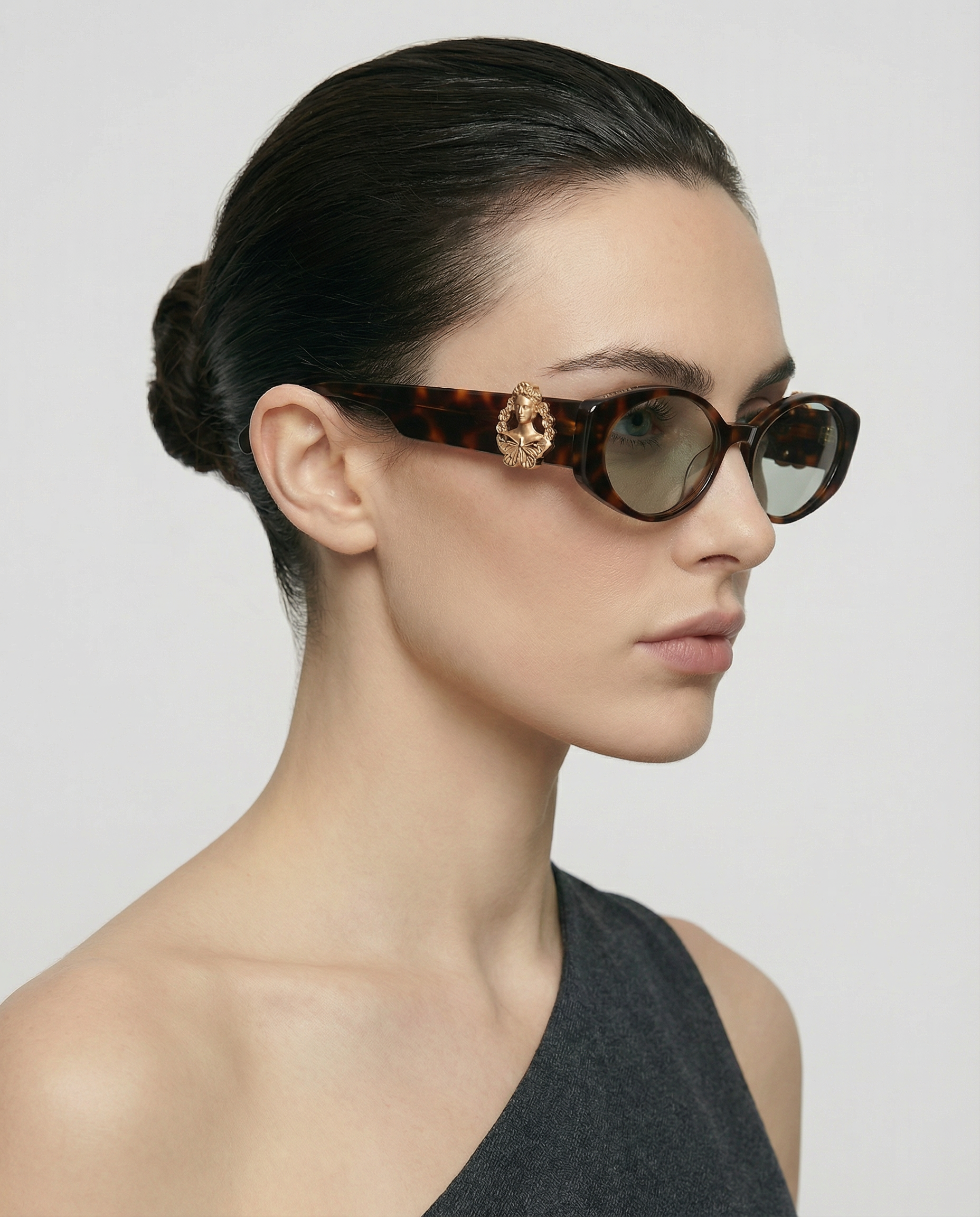 A woman with dark hair in a low bun models the For Art's Sake® Frida Portrait cateye tortoiseshell sunglasses with gold detail and a one-shoulder dark gray top, set against a plain light background.