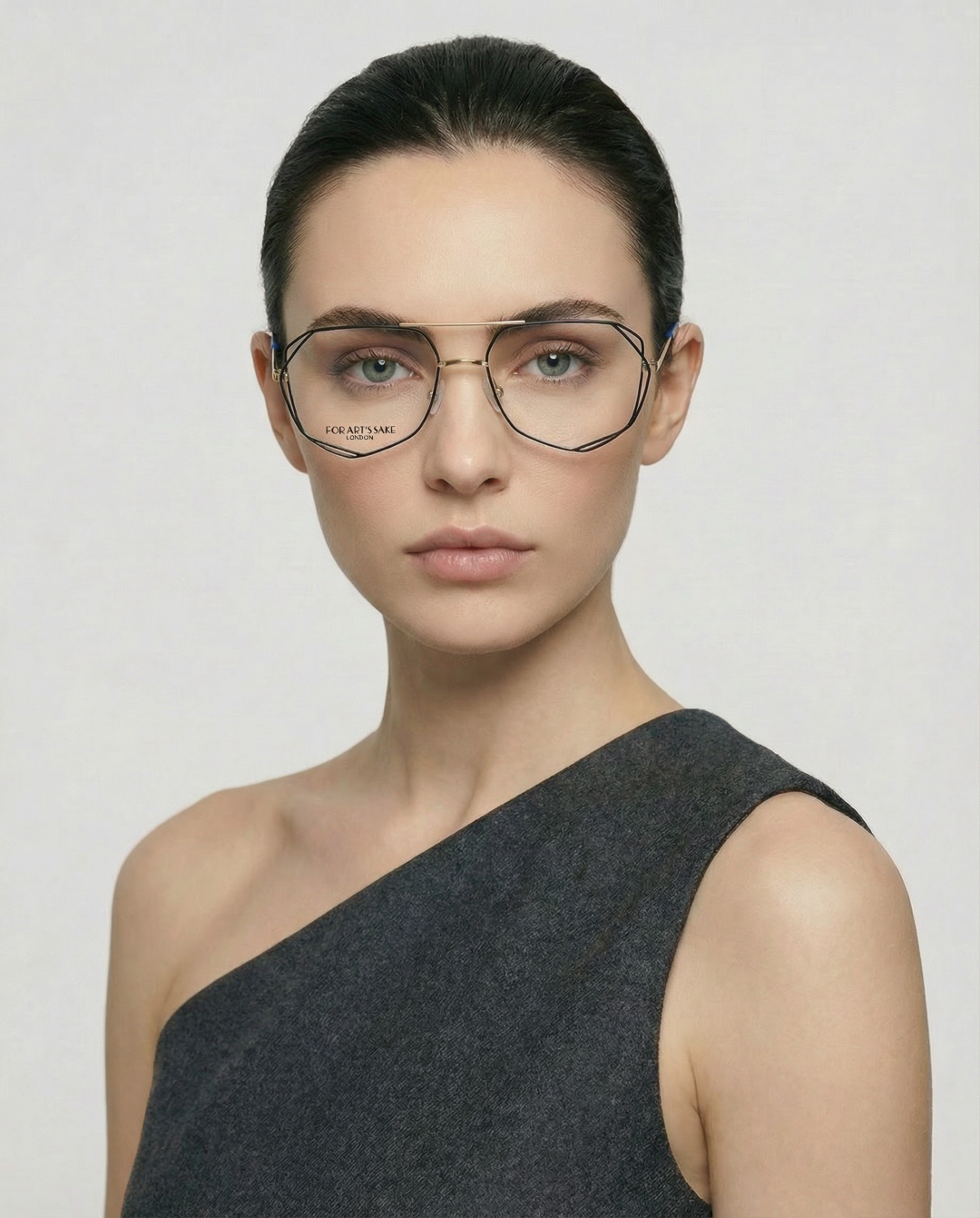 A woman with dark hair pulled back wears oversized Genius Two stainless steel glasses by For Art's Sake®, featuring jade stone nose pads, and a one-shoulder dark grey top, posing against a plain light background with a neutral expression.