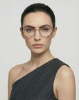 A woman with dark hair pulled back wears oversized Genius Two stainless steel glasses by For Art's Sake®, featuring jade stone nose pads, and a one-shoulder dark grey top, posing against a plain light background with a neutral expression.