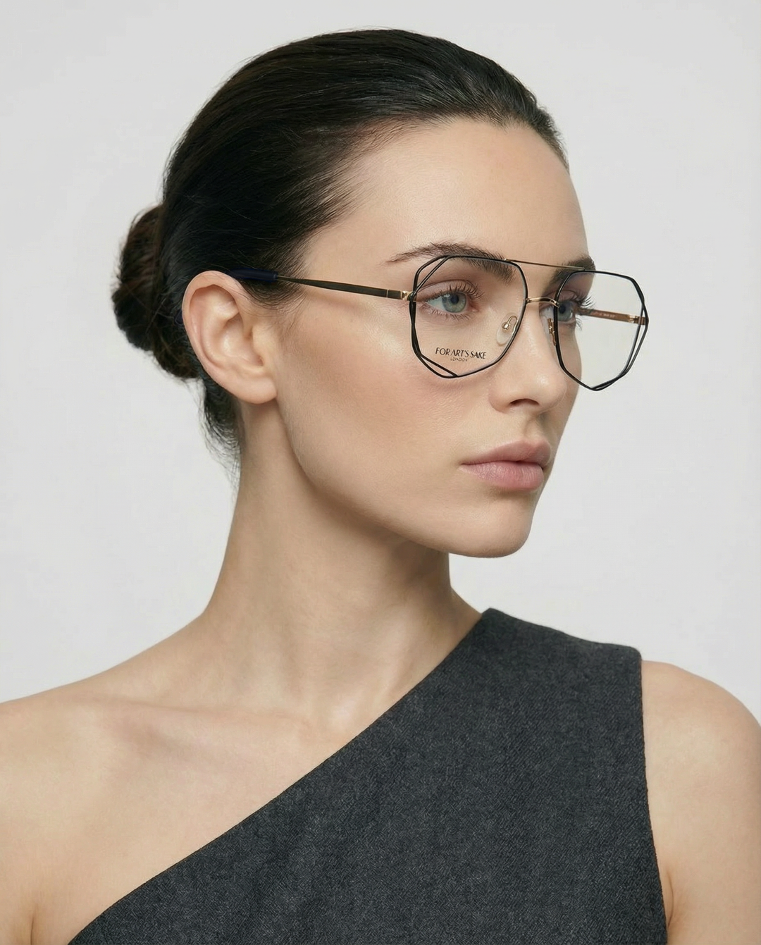 A woman with dark hair tied back, wearing For Art’s Sake® Genius Two geometric stainless steel glasses with jade stone nose pads and a one-shoulder dark grey top, looks to the side against a plain light background.
