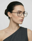 A woman with dark hair tied back, wearing For Art’s Sake® Genius Two geometric stainless steel glasses with jade stone nose pads and a one-shoulder dark grey top, looks to the side against a plain light background.