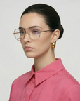 A woman with light skin and dark hair in a low bun wears Genius Two geometric stainless steel glasses by For Art's Sake®, large gold hoops, and a bright pink collared shirt against a plain light background.