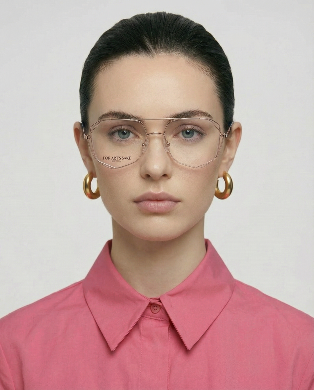 A woman with dark hair in a neat bun wears gold hoop earrings, a buttoned-up pink shirt, and Genius Two unisex opticals by For Art's Sake®, facing forward with a neutral expression against a plain background.