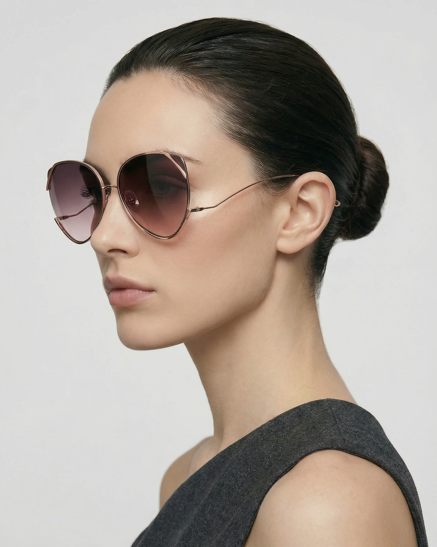 A woman with dark hair in a sleek bun wears For Art's Sake® Wonderland sunglasses—large, round frames with pink-tinted lenses and jadestone nose pads—paired with a sleeveless grey top, gazing left against a plain light background.