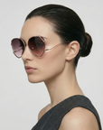 A woman with dark hair in a sleek bun wears For Art's Sake® Wonderland sunglasses—large, round frames with pink-tinted lenses and jadestone nose pads—paired with a sleeveless grey top, gazing left against a plain light background.