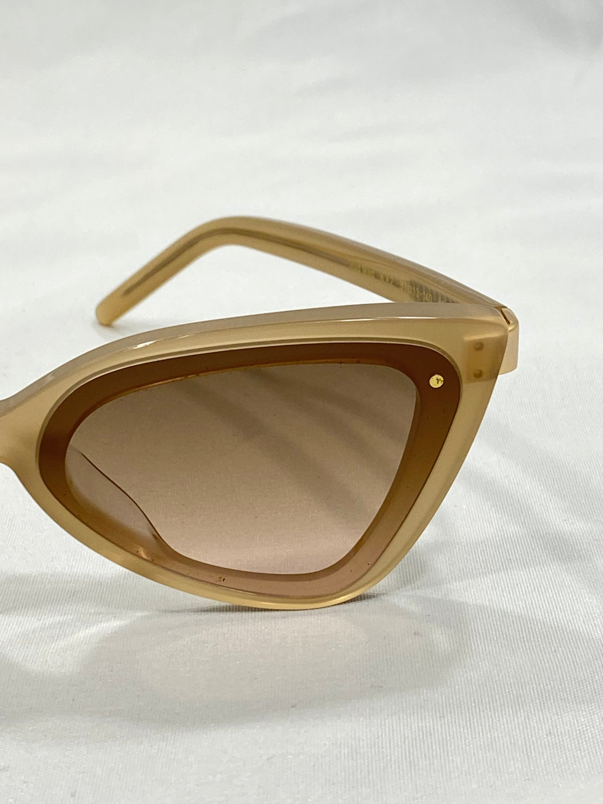 A close-up of For Art's Sake® Mad Mad cat-eye sunglasses in a tan color with slightly tinted lenses. The acetate frames boast a sleek, glossy finish and feature a small metallic detail on the corner. The background is a neutral white fabric, evoking the elegance of Fashion Week.