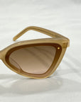 A close-up of For Art's Sake® Mad Mad cat-eye sunglasses in a tan color with slightly tinted lenses. The acetate frames boast a sleek, glossy finish and feature a small metallic detail on the corner. The background is a neutral white fabric, evoking the elegance of Fashion Week.