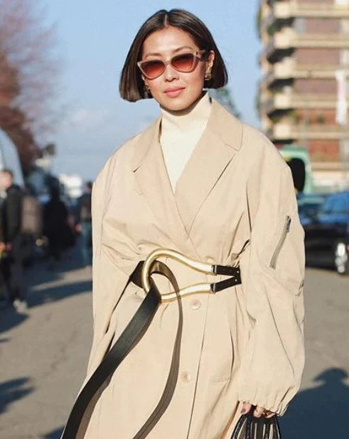 A person with short hair and stylish Mad Mad cat-eye sunglasses from For Art's Sake® is wearing a beige trench coat with a wide belt and a white turtleneck. They are walking down a street with a tall building and blurred figures in the background. The sky is clear and sunny.