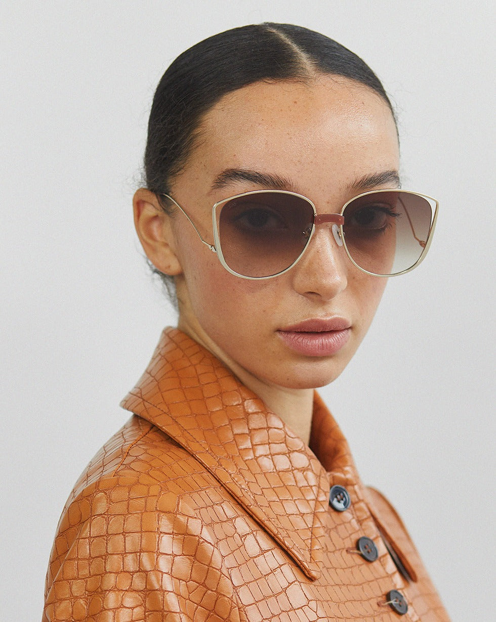 A person with dark, slicked-back hair wears large, stylish Spacie gradient sunglasses from For Art's Sake®, which feature jadestone nosepads and 18-karat gold plating. They look towards the camera with a neutral expression against a plain, light background.