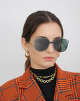 A woman with brown hair is wearing the Spacie by For Art's Sake® – large, dark, handmade sunglasses with jadestone nosepads. She complements them with a gold chain necklace, hoop earrings, and a black turtleneck beneath an orange and black plaid blazer. She stands against a plain white background, looking slightly to the side with a neutral expression.