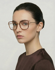 A woman with fair skin and dark hair pulled back wears For Art's Sake® Lace eyeglasses—large frames in brown acetate—and a dark brown top, gazing slightly to the side with a neutral expression against a plain background.
