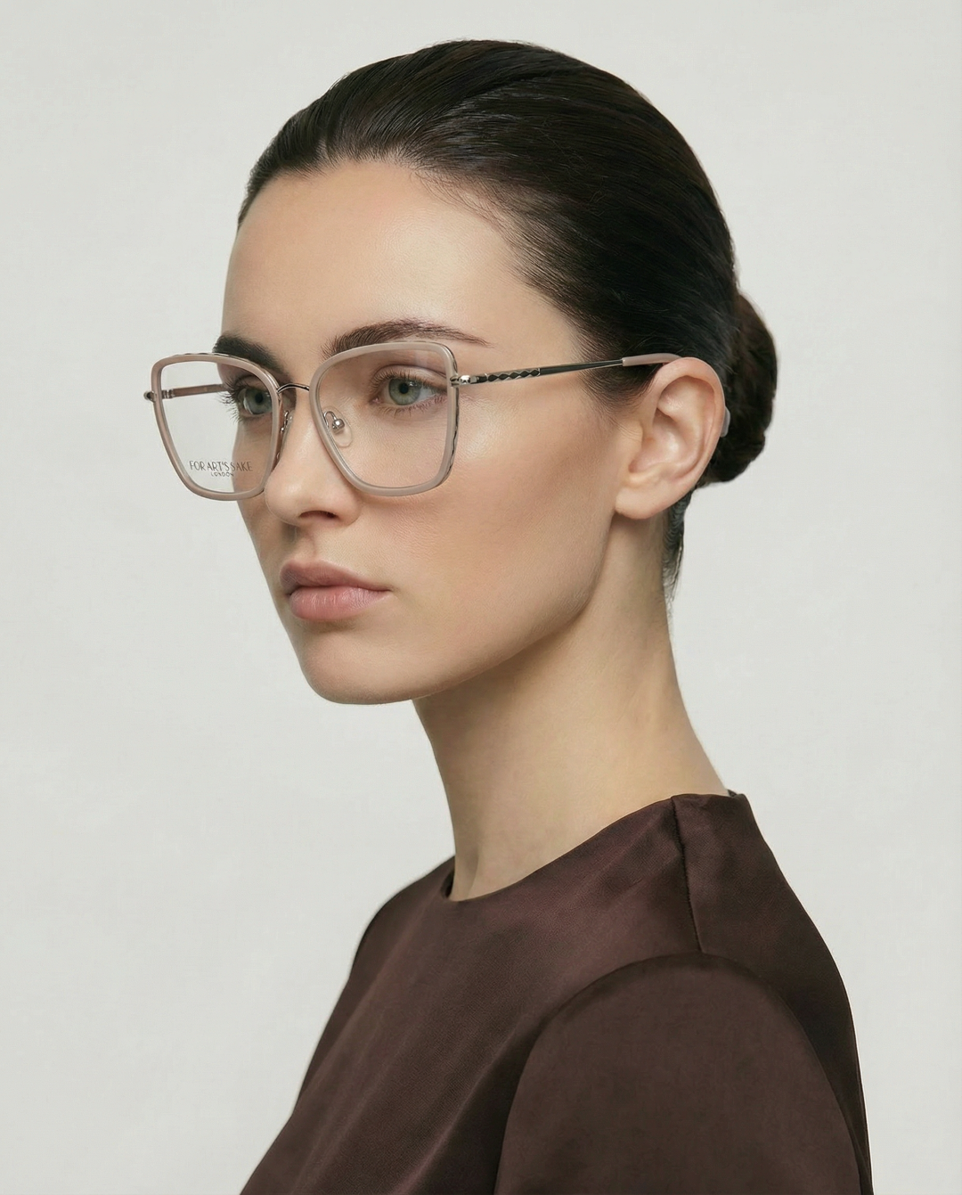 A woman with fair skin and dark hair wears For Art's Sake® Lace square acetate frames with clear jadestone nosepads and a brown top, gazing slightly to the side against a plain light background.