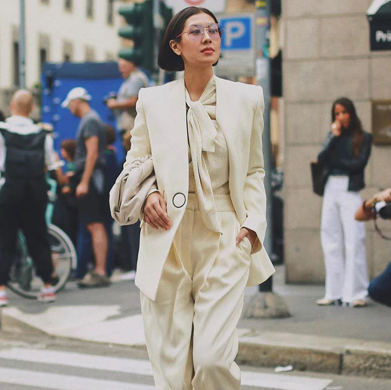 A stylish individual walks confidently down a city street wearing an off-white pantsuit with a matching tie-neck blouse. They carry a bag under one arm and sport UV-protected aviator shades from For Art's Sake® called Explorer Pink. In the background, people are engaged in various activities near a busy intersection.
