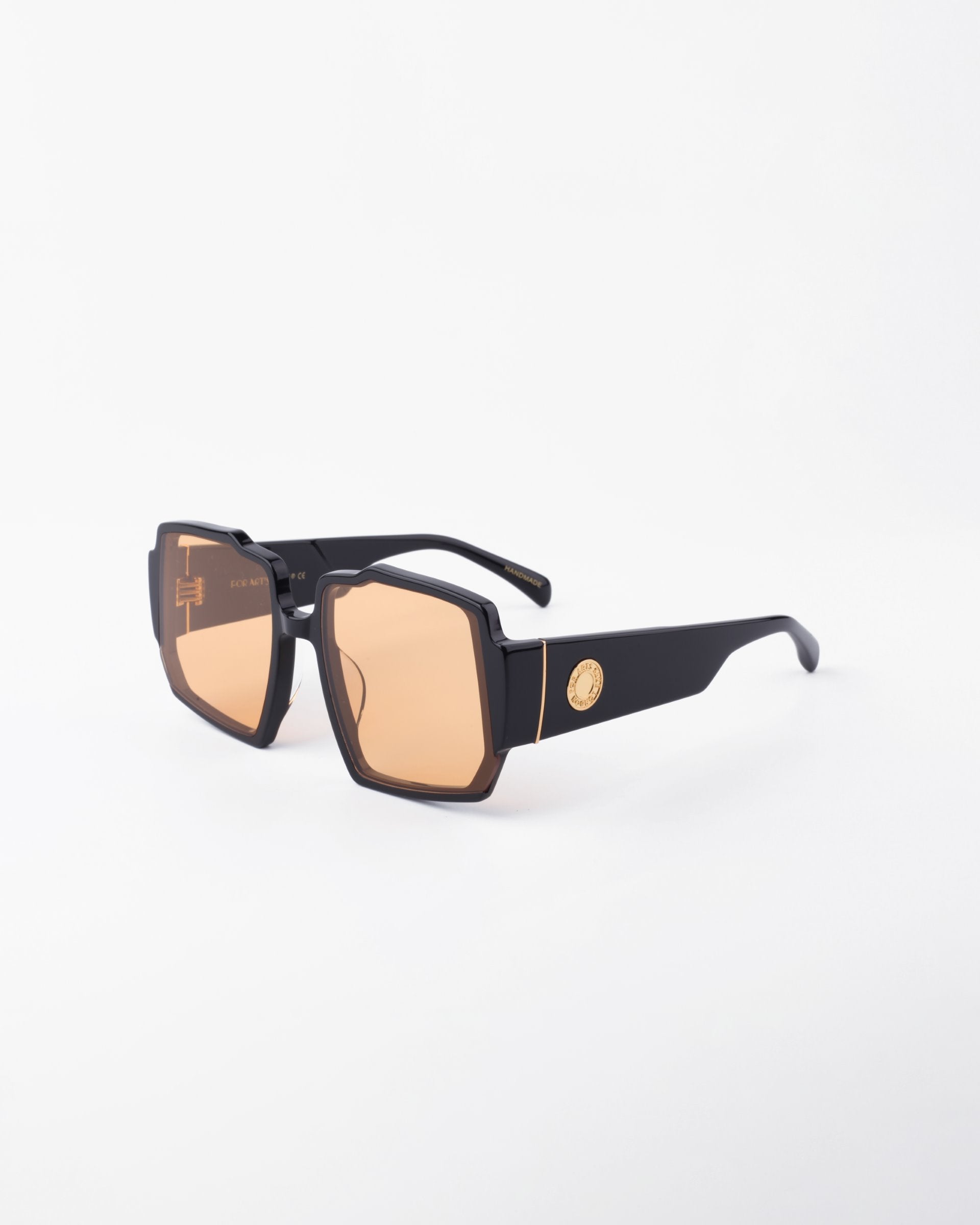 A pair of stylish black For Art's Sake® Moritz sunglasses with large, geometric, orange-tinted UV-protected lenses sits against a plain white background. The sunglasses feature a chunky acetate frame and thick arms adorned with a small, gold-colored emblem near the hinge.