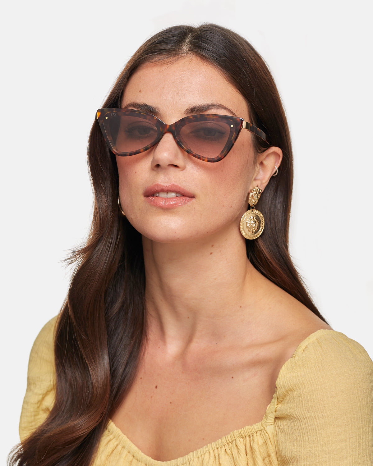 A woman with long, dark hair is wearing oversized, brown cat-eye sunglasses with acetate frames and large gold earrings. She is dressed in a light yellow off-shoulder top and looking slightly to the side against a plain white background, wearing Mad Mad Champagne by For Art's Sake®.