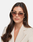 A woman with long brown hair, wearing a beige blazer and angular Mad Mad sunglasses by For Art's Sake®, gazes ahead with a neutral expression. Her light-brown acetate frame sunglasses feature a gold accent on the temples. She also wears gold hoop earrings, with a plain white background behind her.