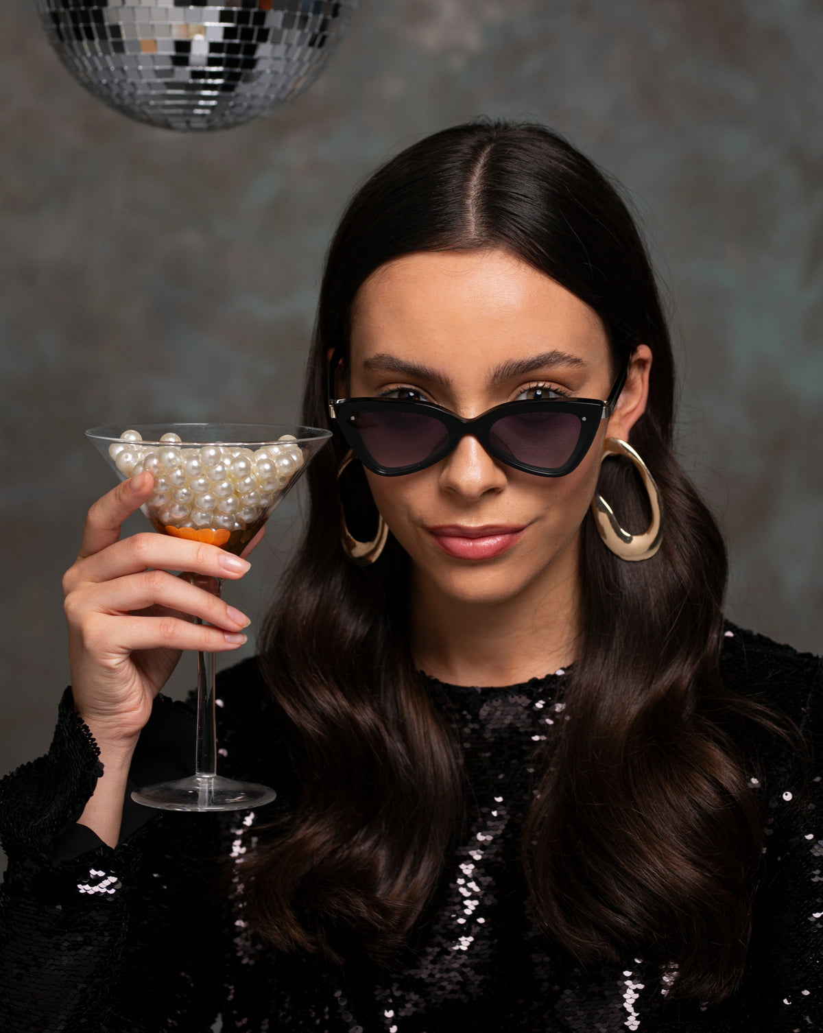 A woman with long dark hair and large hoop earrings is wearing For Art's Sake® Mad Mad cat-eye sunglasses with acetate frames and a black sequined top. She holds a martini glass filled with pearls, while a disco ball hangs above her against a dark textured background.