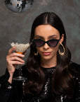 A woman with long dark hair and large hoop earrings is wearing For Art's Sake® Mad Mad cat-eye sunglasses with acetate frames and a black sequined top. She holds a martini glass filled with pearls, while a disco ball hangs above her against a dark textured background.