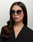 A woman with long, dark hair wears oversized, hexagonal For Art’s Sake® Moma shades with stainless steel frames and a black blazer. She has hoop earrings and a neutral expression, posing against a plain, light background.