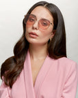 A woman with long brown hair is wearing For Art's Sake® Moma shades with stainless steel frames and gold earrings. She is dressed in a light pink blazer and is facing the camera with a neutral expression against a plain white background. Her sunglasses are 100% UVA and UVB-protected.
