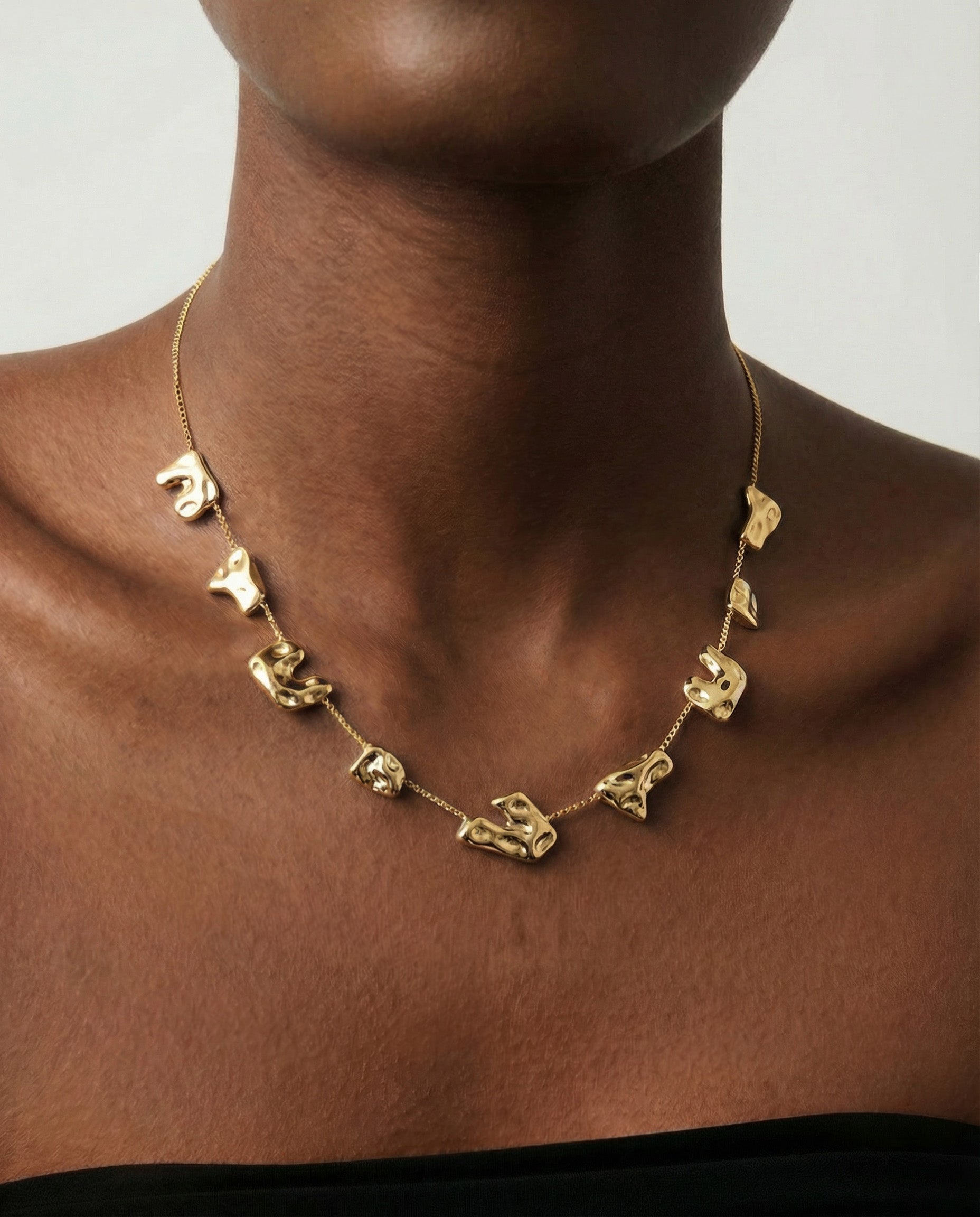 Someone wears The Forms Necklace by For Art's Sake®, a modern gold piece with abstract, mask-shaped charms. It rests on their bare upper chest above a black strapless top, set against a plain light background.