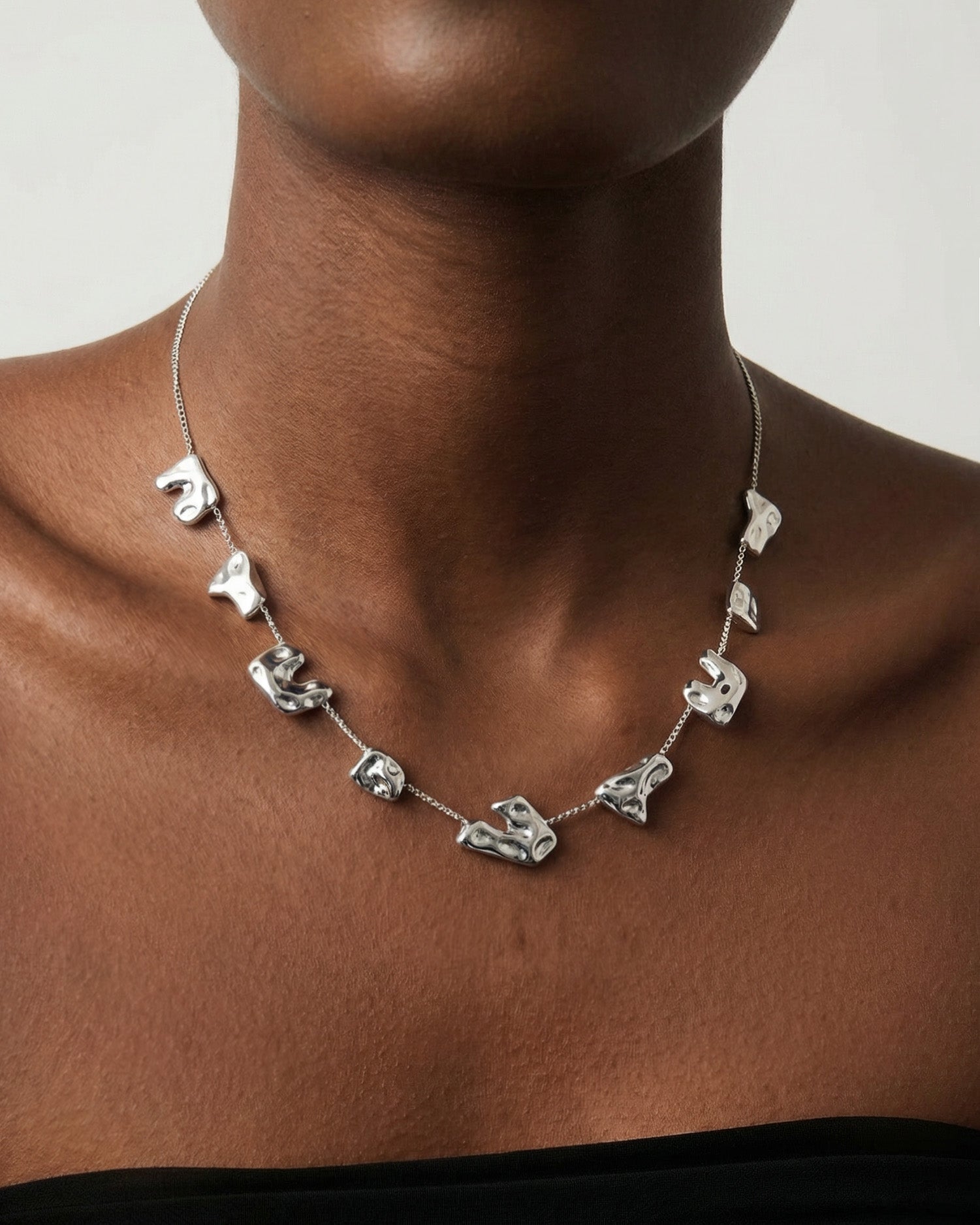 Wearing For Art's Sake® The Forms Necklace—a silver organic metal piece with evenly spaced abstract shapes—they pose in a black strapless top against a plain background, highlighting this contemporary jewelry.
