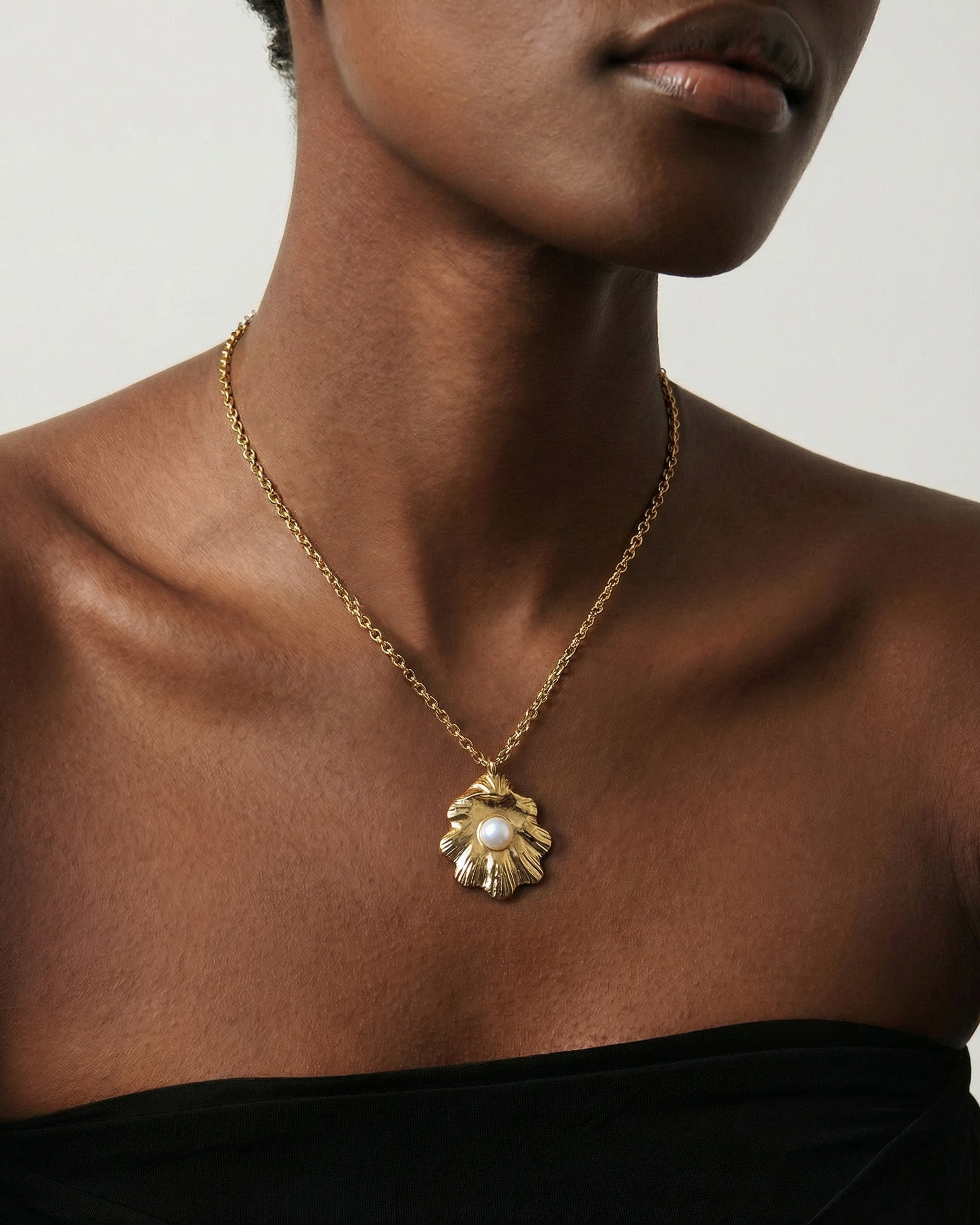 A woman models The Shell Pendant by For Art’s Sake®—a gold sculptural sea shell necklace with a central freshwater pearl—styled with a black strapless top against a neutral backdrop to highlight its elegance.