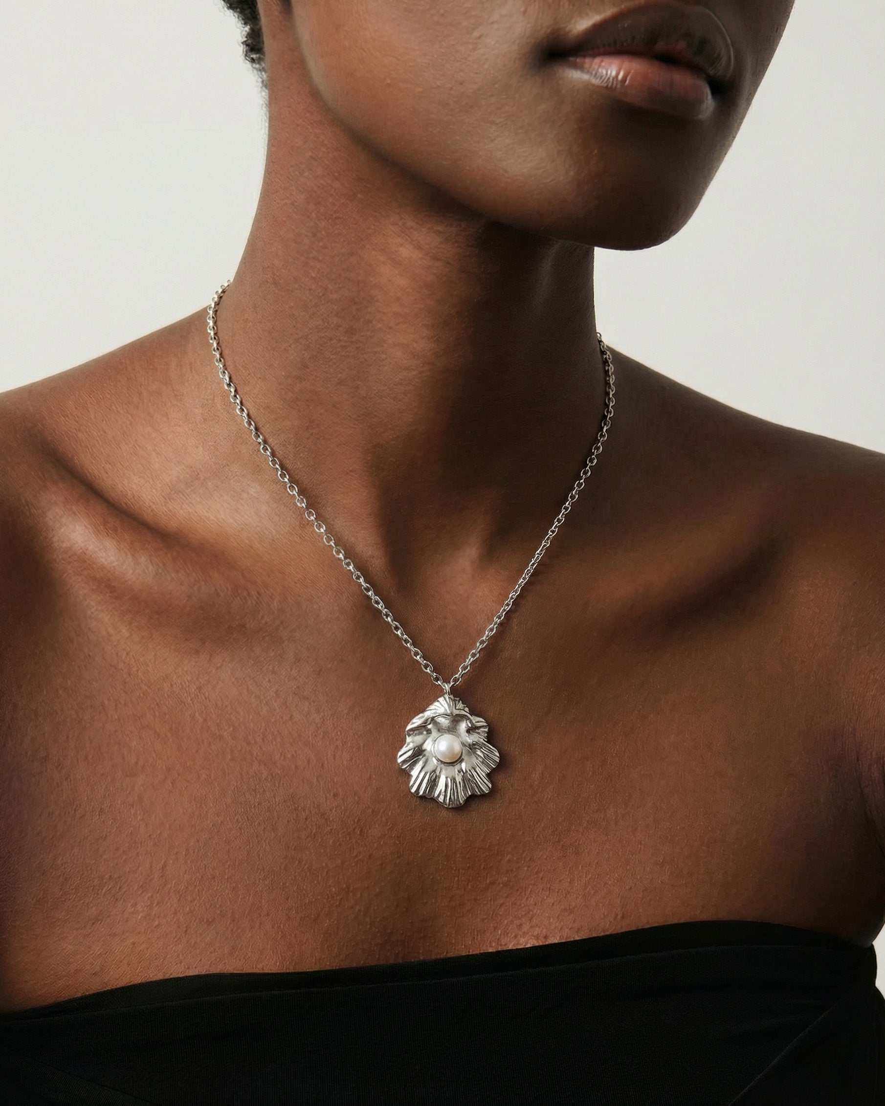 A model in a strapless black top highlights The Shell Pendant by For Art's Sake®—a sea shell necklace featuring a standout freshwater pearl—against a plain background.