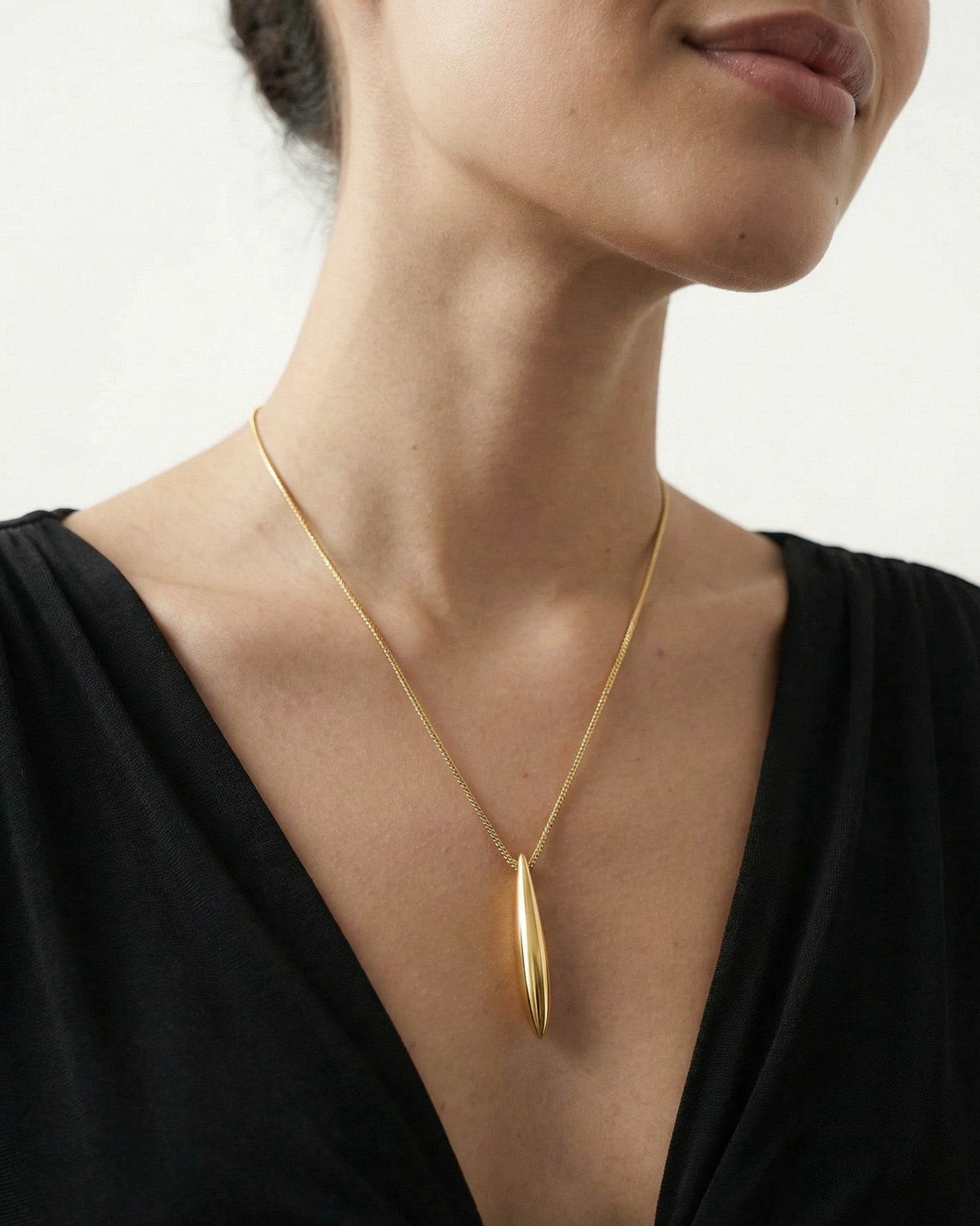 A woman wears The Droplets Pendant Necklace by For Art's Sake®—a gold modern necklace with a long, metal droplet pendant on a delicate chain—paired with a black V-neck top against a plain light background.