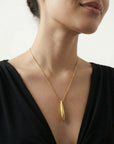 A woman wears The Droplets Pendant Necklace by For Art's Sake®—a gold modern necklace with a long, metal droplet pendant on a delicate chain—paired with a black V-neck top against a plain light background.