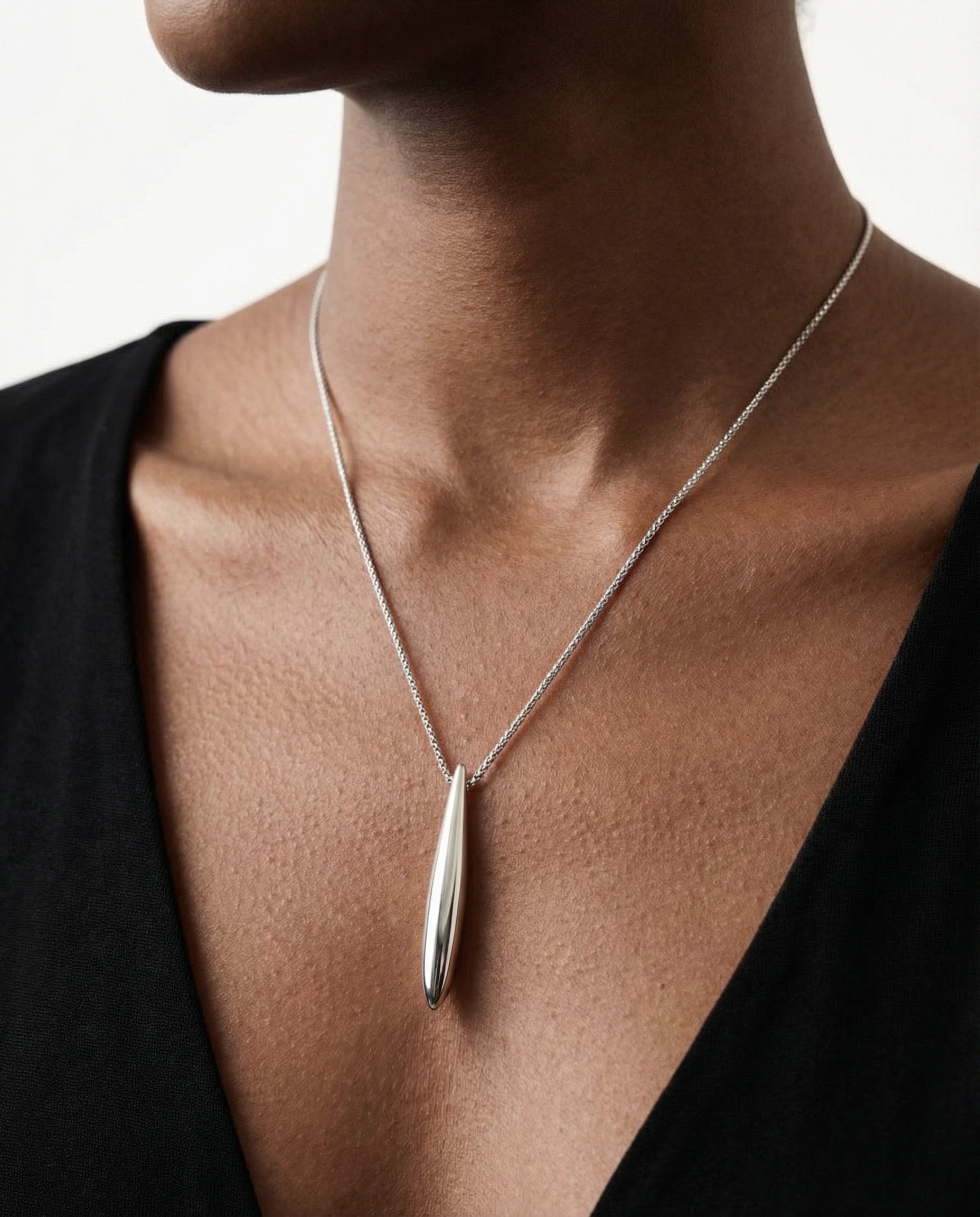 A close-up of a person with medium-brown skin in a black V-neck top wearing For Art's Sake® The Droplets Pendant Necklace, featuring a sleek silver elongated metal droplet pendant on a delicate chain.