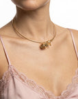 A woman models The Rose Choker by For Art’s Sake®—a delicate gold necklace with a sculpted rose and leaf charm—paired with a pink lace-trimmed camisole. Only her neck, shoulders, and upper chest are seen against a white background.
