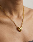 A close-up of a person wearing For Art's Sake® The Sculpted Heart Necklace, featuring a chunky gold chain and sculptural heart pendant, against bare skin and a strapless dark top.