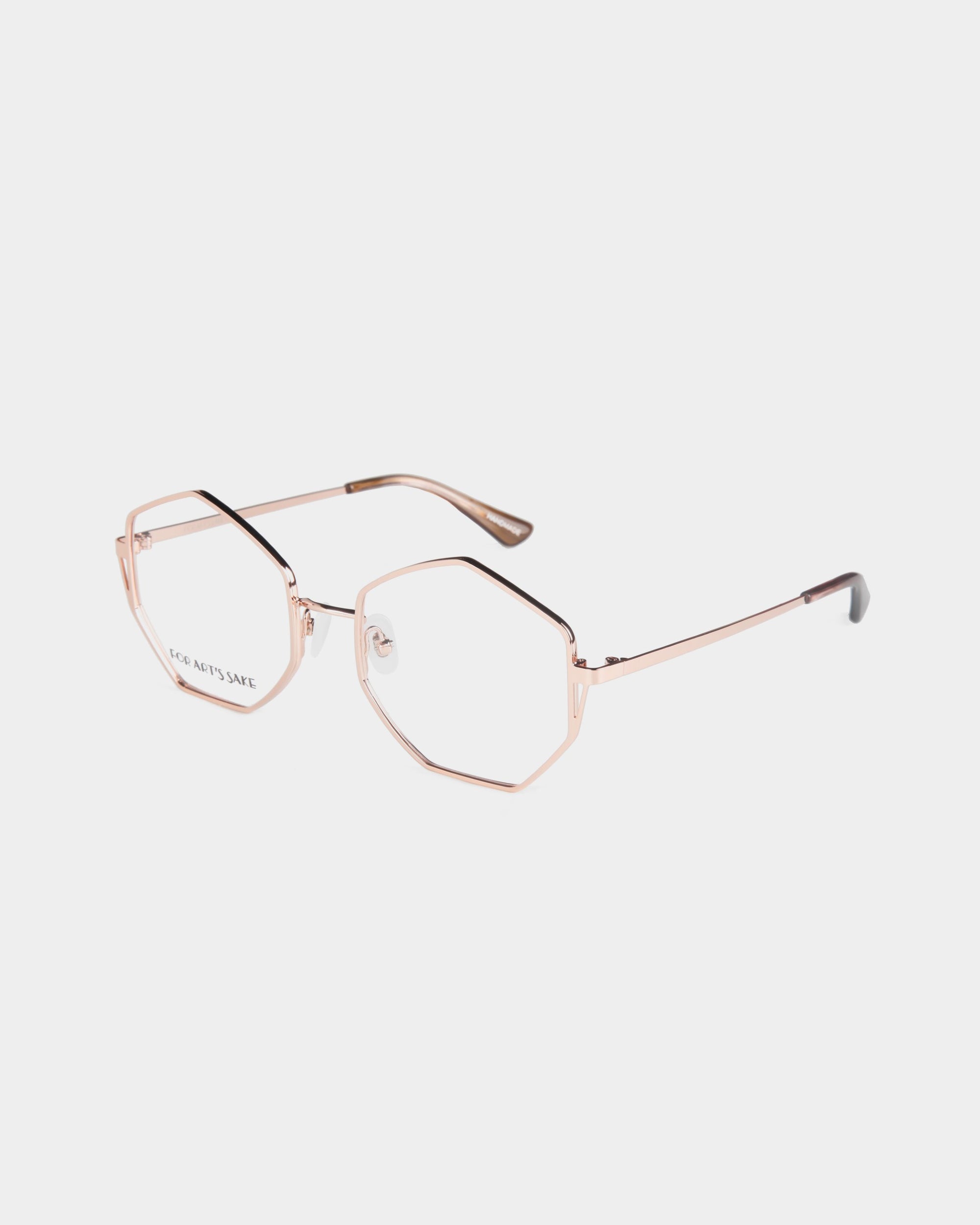 A pair of rose gold For Art's Sake® Antidote glasses featuring heptagon-shaped clear lenses. The minimalist design boasts thin metal arms with black tips at the ends, and the glasses are positioned on a white background. Jade gemstone nose pads add a touch of luxury to this elegant accessory.