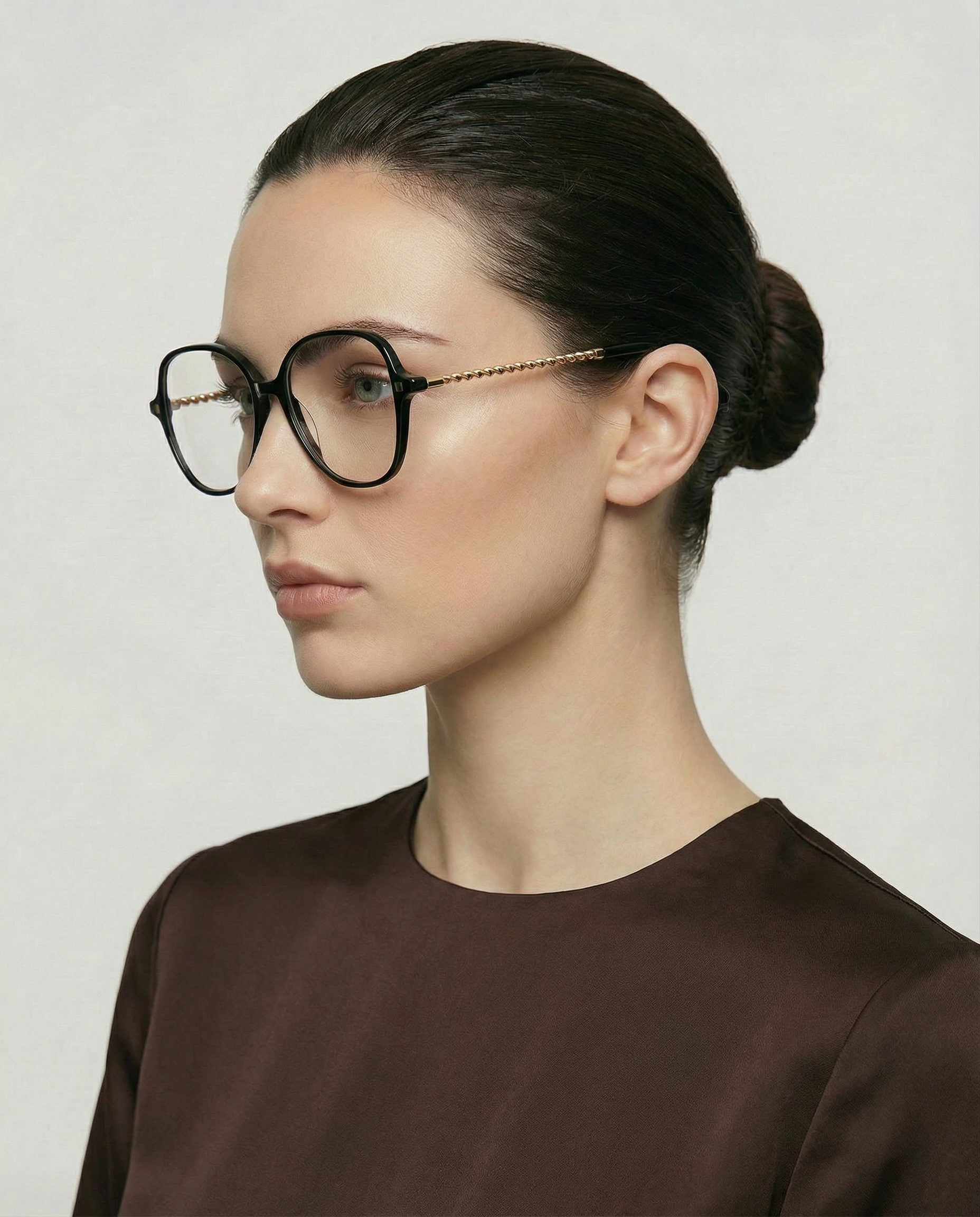 A woman with fair skin and dark hair in a low bun wears For Art's Sake® Dumpling large, round black acetate frames with gold-plated arms and a decorative gold chain. She is dressed in a dark brown top and faces left against a plain background.