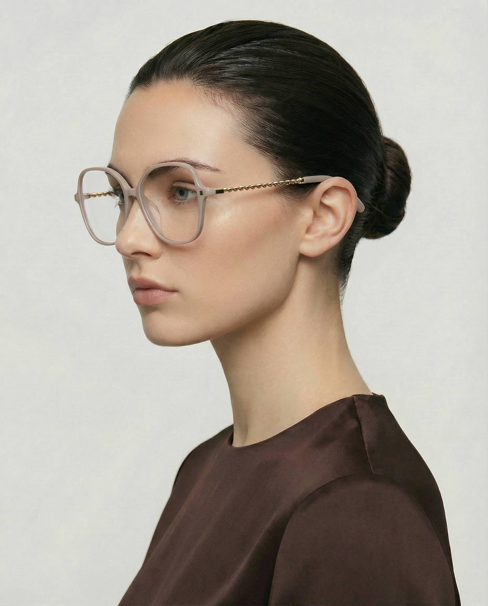 A woman with dark hair in a bun wears For Art's Sake® Dumpling oversized clear acetate frames with gold-plated arms and chain details, paired with a brown top, looking left against a light background.