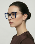 A woman with dark hair in a low bun wears For Art's Sake® Dion cat-eye blue light filter glasses and a brown top, posing in profile against a plain light background.