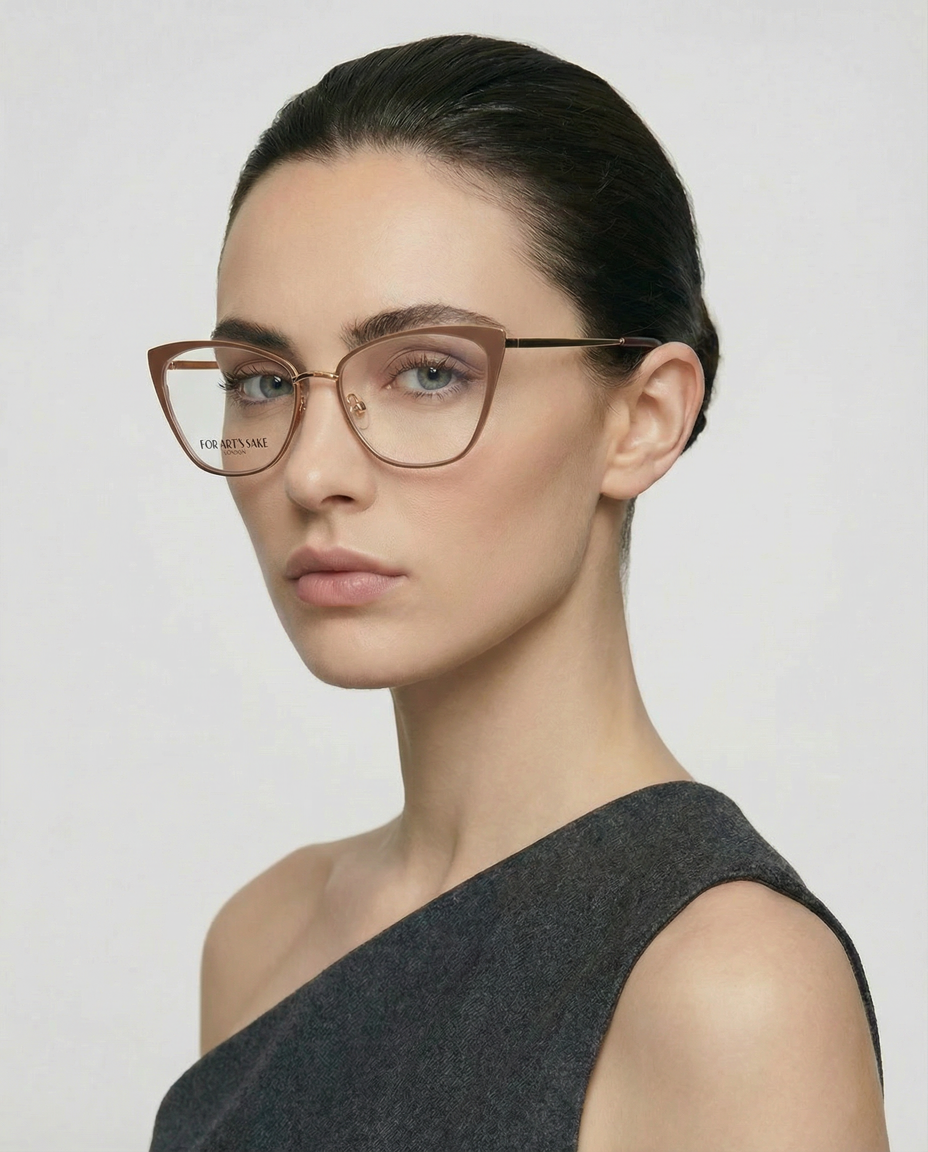 A woman with dark hair tied back wears For Art's Sake® Stella Two cat-eye glasses and a one-shoulder dark gray top, looking neutrally at the camera against a plain light background.